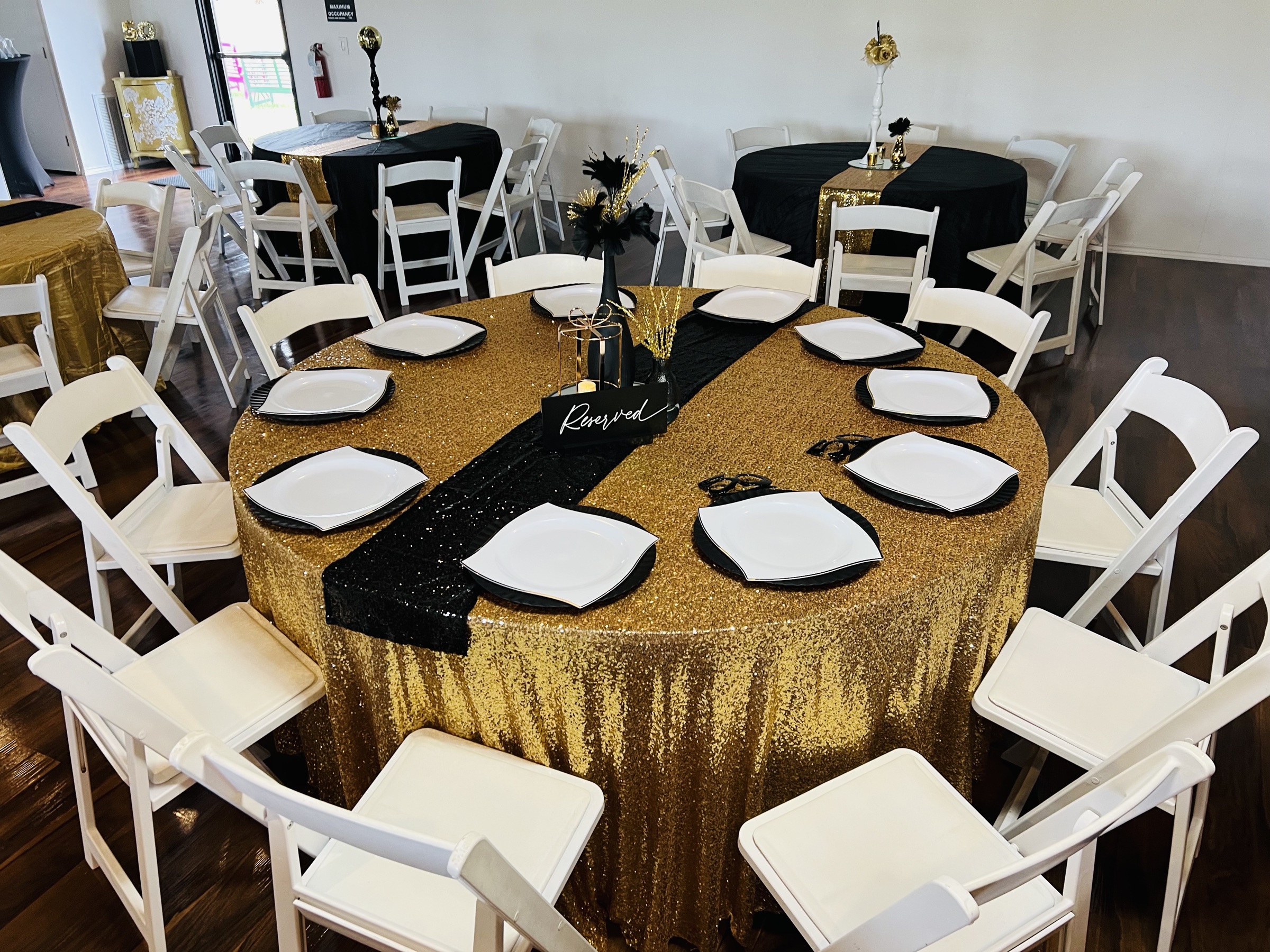 A black-and-gold gala table - gold sequin runner, black napkins, white plates with crystal-trimmed chargers, white folding chairs.